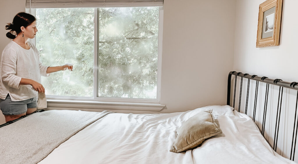 woman spraying cleaner on a window next to a bed. 